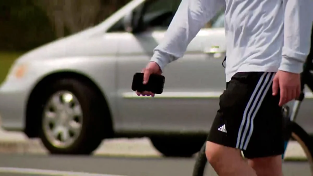 Picture of Caucasian male person holding a cellphone walking past a white vehicle.
