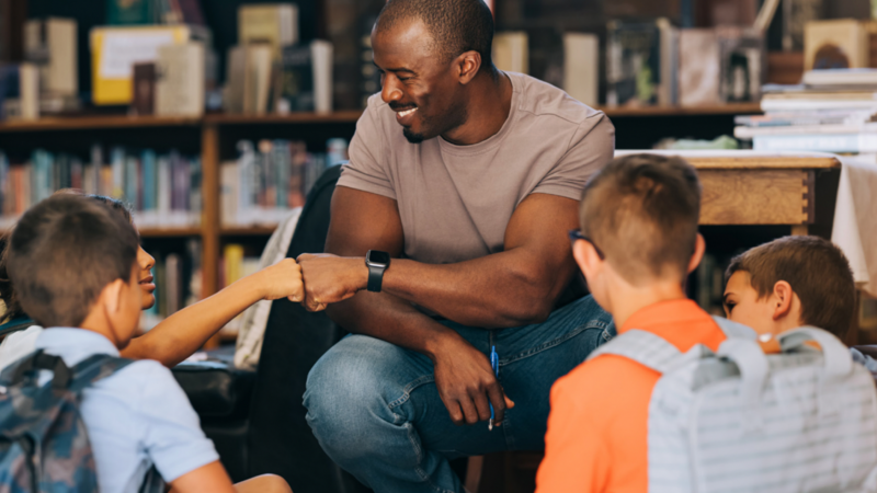 Adult mentor seated with a small group of students in a library, engaging in a friendly discussion and exchanging a fist bump with one child.