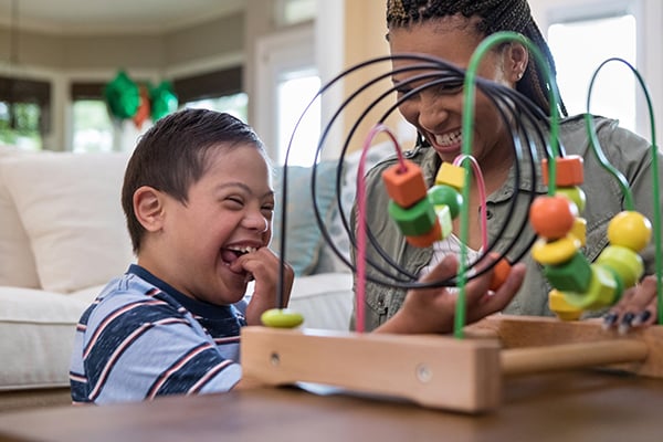 mother and son with Down syndrome laughing and playing with bead toy in living room.